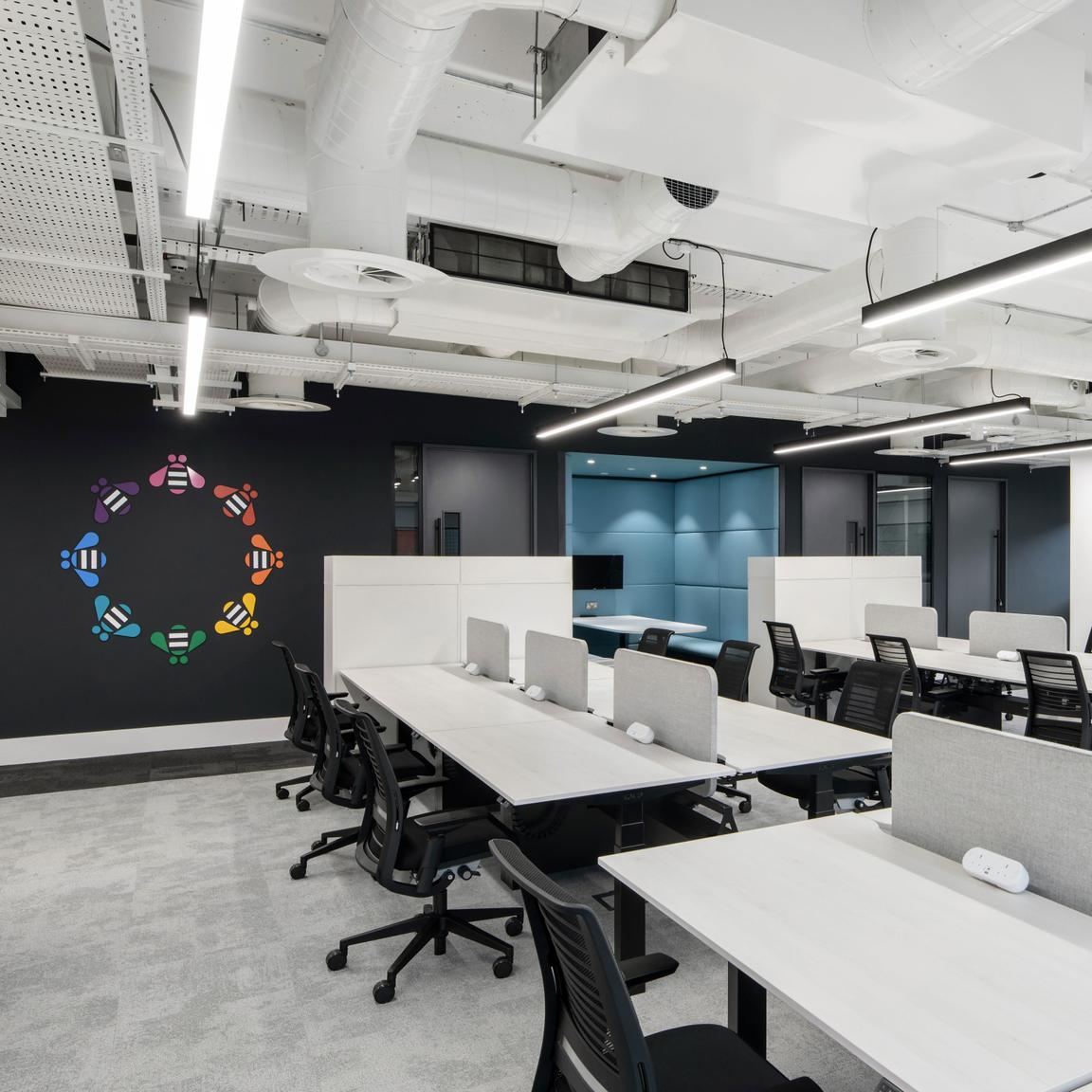 Open plan floor with height-adjustable desks in screened pairs and a glass meeting booth beyond